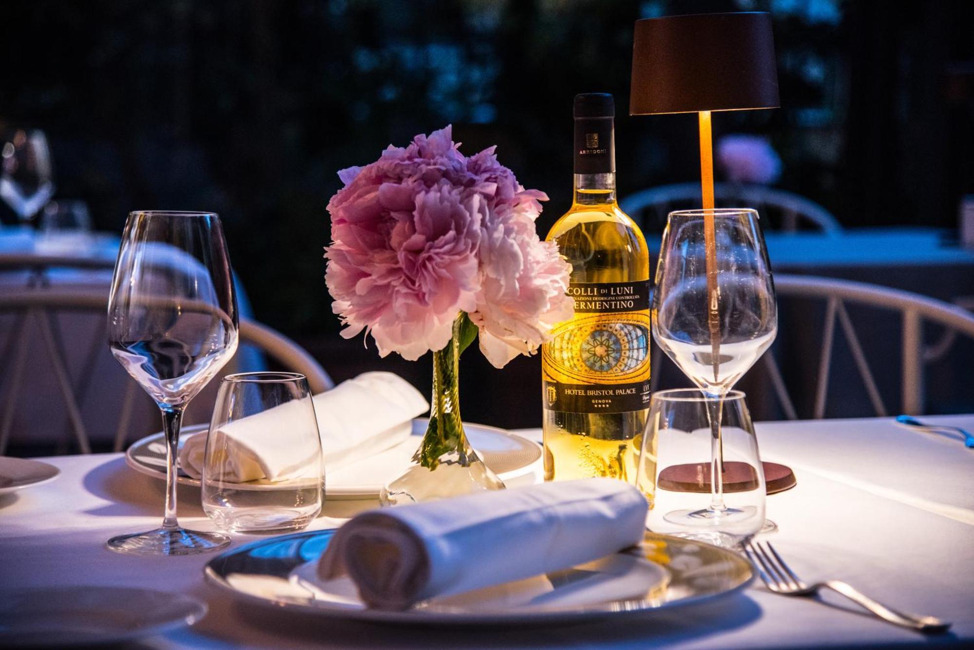 Hotel Bristol Palace Генуя Экстерьер фото The photo shows a beautifully set dining table with an elegant ambiance. There is a vase with a pink flower arrangement at the center, adding a touch of color and freshness. A bottle of white wine is also prominently displayed next to the flowers. Th