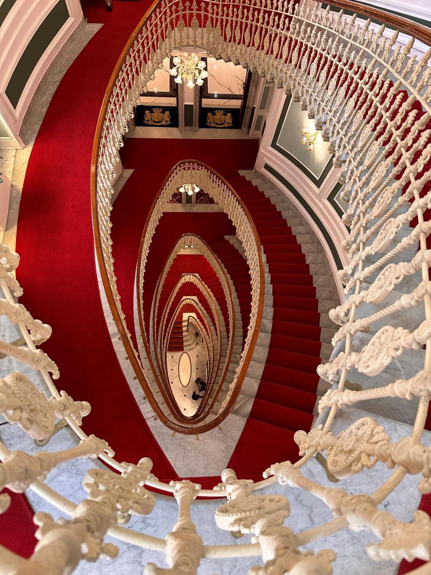 Hotel Bristol Palace Генуя Экстерьер фото The photo depicts a spiraling staircase viewed from above. The staircase features ornate, white railings and a striking red carpet that runs down the steps. The spiral design creates a visual effect of depth, leading the eye downwards. The surroundin