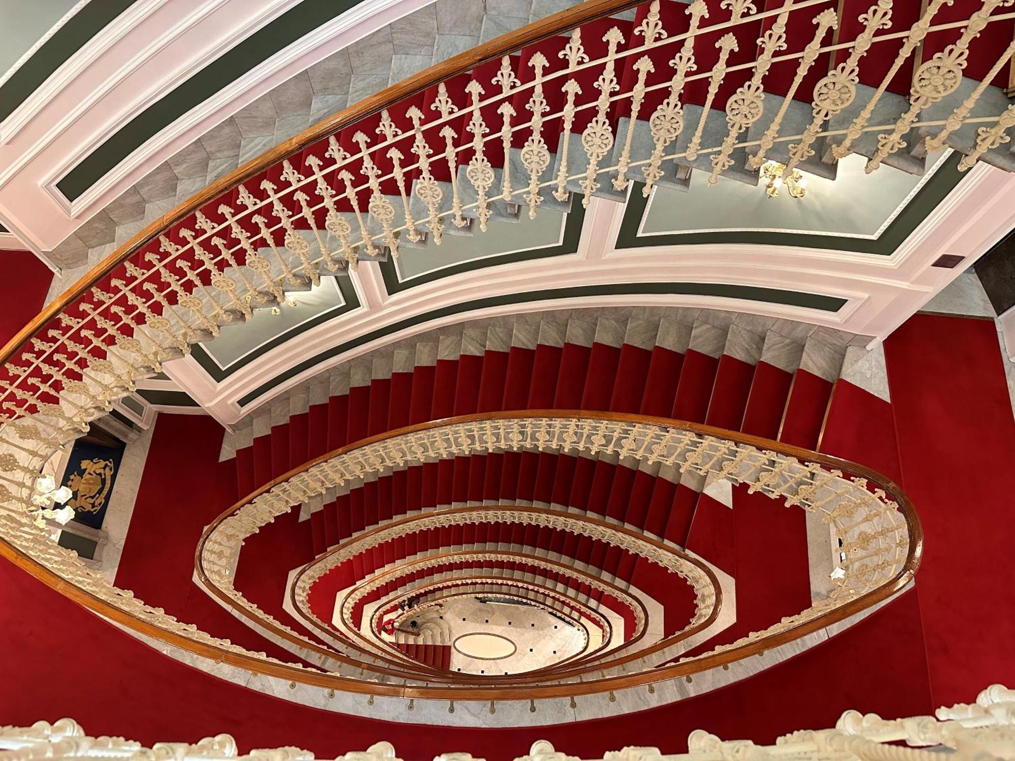 Hotel Bristol Palace Генуя Экстерьер фото The photo shows a grand, spiral staircase viewed from above. The staircase features elegant white railings with intricate designs and is surrounded by a rich red carpet. The walls have a classic architectural style, with decorative elements in soft c
