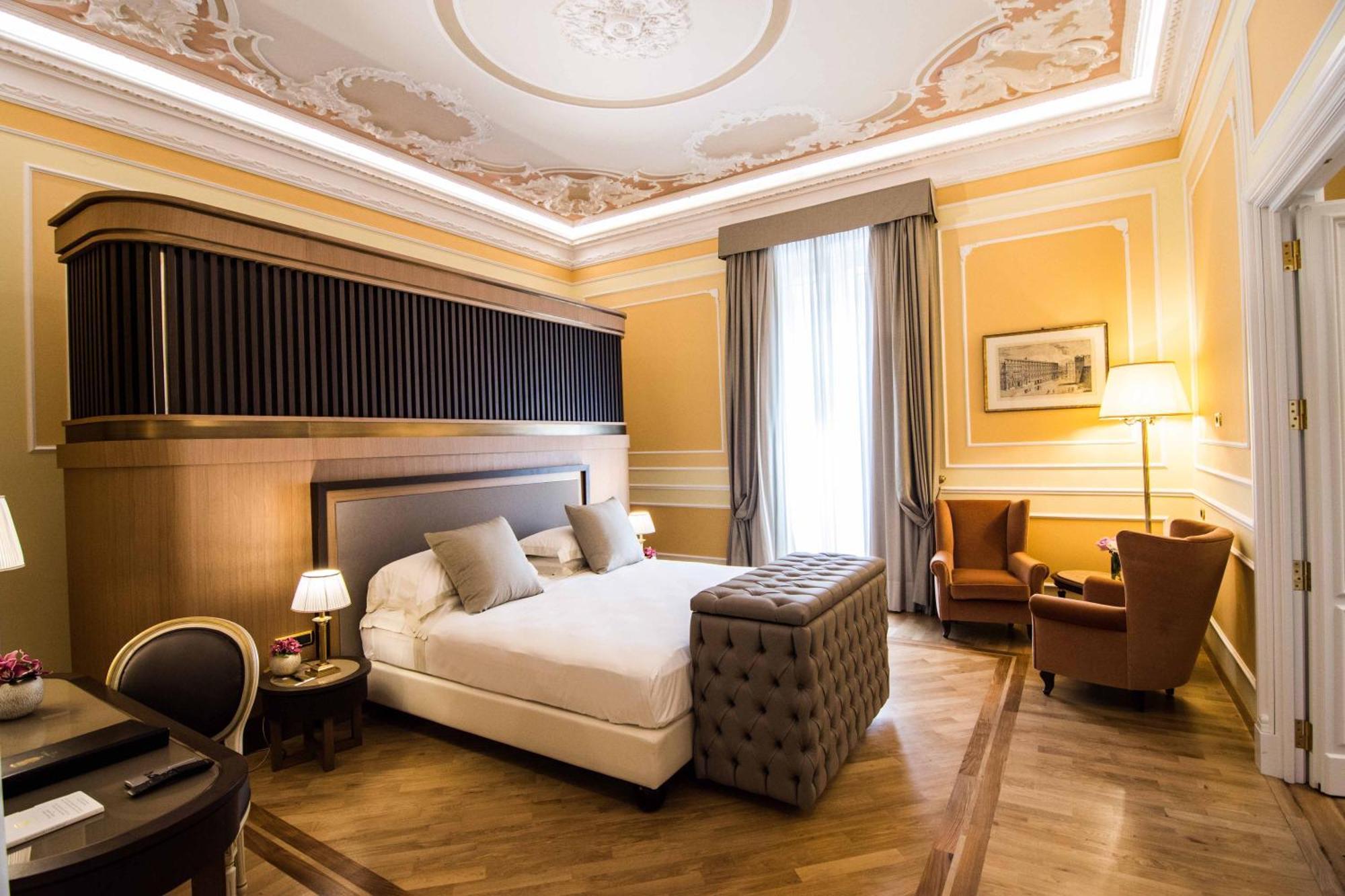 Hotel Bristol Palace Генуя Экстерьер фото The photo shows an elegantly designed hotel room. It features a large bed with plush pillows, and a tufted ottoman at the foot of the bed. The walls are painted in a warm yellow tone, adorned with decorative molding. A pair of tall windows are draped