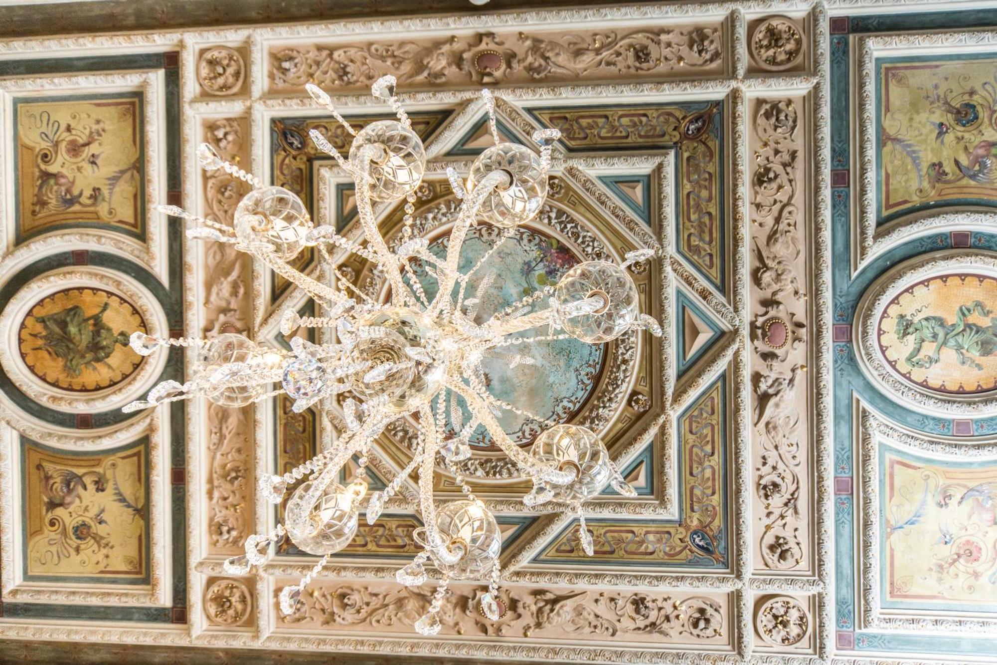 Hotel Bristol Palace Генуя Номер фото The image depicts an ornate ceiling featuring a large chandelier made of crystal. The chandelier hangs from a richly detailed ceiling adorned with intricate designs, including decorative molding and colorful frescoes. The artwork includes circular mo